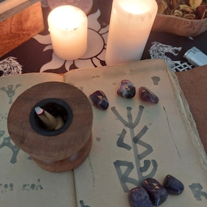 May include: A mystical scene featuring lit candles, a wooden incense holder with an incense stick, and several purple rune stones. The stones are placed on an open book with a rune symbol. Dried herbs and a wooden box are also visible.