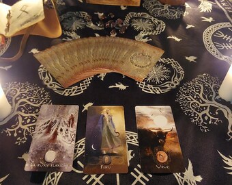May include: A collection of tarot cards with illustrations, including one with the text "LA RUNE BLANCHE". The cards are arranged on a black cloth with white patterns, alongside a fan, candles, and a wooden box.