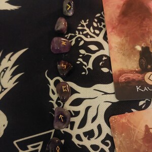 Peut inclure: Pierres de runes en améthyste avec des symboles dorés, disposées verticalement. Les pierres sont sur un tissu noir avec un motif blanc. Une carte de tarot avec une image brun-rouge est partiellement visible.