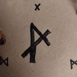 Peut inclure: Gros plan sur une surface beige avec plusieurs symboles runiques noirs. Le symbole central est une ligne diagonale croisée par une autre, ressemblant à la lettre 'A'. D'autres symboles incluent un 'X' et un 'X' miroir.