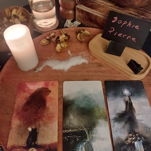 May include: A still life arrangement featuring three tarot cards with illustrations and text, a lit candle, a small bottle, and a wooden tray with a handwritten name. The cards are labeled "Kaunan", "Laukaz", and "Isaz".