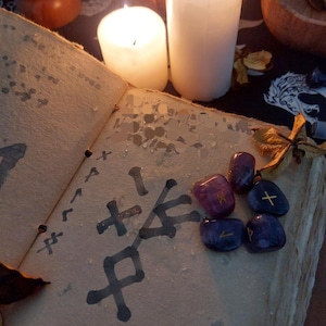 Peut inclure: Un livre ouvert avec des symboles manuscrits et une collection de pierres précieuses violettes et bleues. Deux bougies blanches allumées et un fond sombre créent une ambiance mystique.