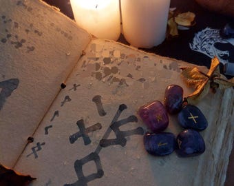 May include: An open book with handwritten symbols and a collection of purple and blue gemstones. Two lit white candles and a dark background create a mystical ambiance. The scene suggests a spiritual or occult theme.