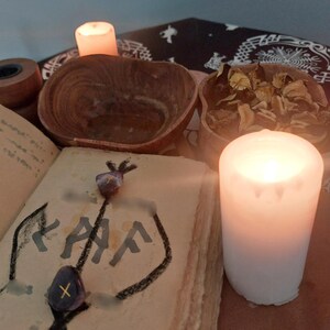 Puede incluir: Una vela blanca encendida junto a un libro abierto con un dibujo y texto. También se ven dos cuencos de madera y una vela pequeña. La escena tiene un tema místico o espiritual.
