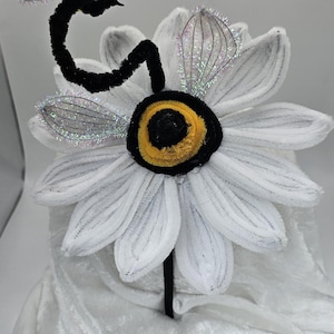 Può includere: Una fascia stravagante con una grande margherita bianca e un centro giallo e nero. Un'ape nera e gialla con ali scintillanti è attaccata. La fascia è nera.