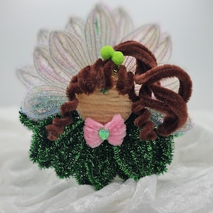 May include: A handcrafted fairy ornament featuring brown pipe cleaner hair, a green tinsel body, and a pink bow. The fairy has iridescent wings and two green pom-poms. This whimsical decoration is ideal for a child's room or as a festive ornament.