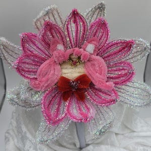 Könnte beinhalten: Ein dekoratives Stirnband mit einem großen, stilisierten Blumenmuster. Die Blume hat rosa Blütenblätter mit silbernen Glitzerakzenten und eine rote Schleife. Die Mitte der Blume hat ein plüschiges rosa Design. Das Stirnband ist weiß.