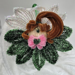 May include: A whimsical handmade ornament featuring a central figure with brown pipe cleaner hair, a pink bow, and a beige face. The figure is surrounded by green tinsel leaves and iridescent wings, perfect for holiday decor.