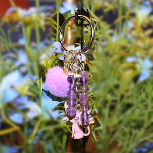 May include: A keychain with a gold-colored ring, featuring a light purple pom-pom, a string of purple beads, and a pink charm of a cartoon cat. The keychain is designed to hold keys or be used as a decorative accessory.