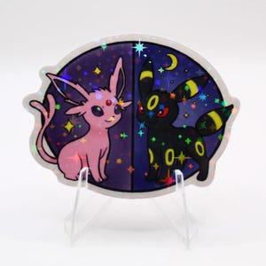 May include: Holographic sticker featuring two cartoon creatures, one pink and one black, set against a starry night sky background. The sticker is oval-shaped and displayed on a clear stand.