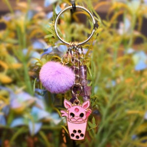 May include: A gold-colored keychain with a pink boba tea charm featuring a cat design, a fluffy purple pom-pom, and purple beads. The keychain is attached to a metal ring, perfect for keys or bags.