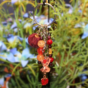 May include: A keychain with a silver ring and multiple charms. The charms include a burgundy pom-pom, orange and red beads, and a cartoon fox pendant with a red flower charm. The background is blurred with green and blue foliage.