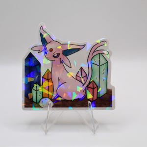 May include: Holographic sticker featuring a pink cartoon creature with large ears and a long tail, surrounded by colorful crystal formations. The sticker has a shiny, iridescent finish and is displayed on a clear stand.