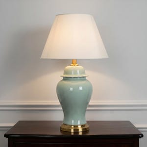 Peut inclure: Une lampe de table avec une base en céramique vert clair en forme de pot à gingembre, dotée d'une finition brillante, d'un col et d'une base circulaire en métal doré. L'abat-jour en tissu blanc diffuse une lumière douce sur la céramique.