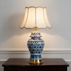 Peut inclure: Une lampe de table avec une base en porcelaine bleu et blanc en forme de pot à gingembre ornée de motifs floraux. La lampe possède un socle en métal doré et un abat-jour en tissu crème à bord festonné et bordure décorative.