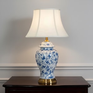 May include: A table lamp with a white lampshade and a blue and white porcelain base. The base features a floral and bird design, with a gold-coloured base and accents. The lamp is placed on a dark wooden surface.