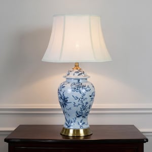 Peut inclure: Une lampe de table dotée d'un abat-jour en tissu blanc et d'une base en céramique ornée d'un motif floral bleu et blanc. La lampe possède un col en métal couleur laiton et une base circulaire assortie, assurant une assise stable.