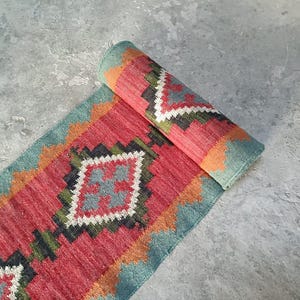 May include: A rectangular woven runner rug featuring a repeating geometric pattern in shades of red, teal, orange, white, and dark green. The rug is partially rolled at one end, displaying a symmetrical diamond motif with contrasting borders.