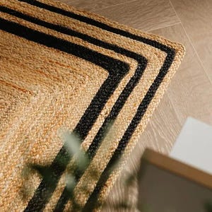 Puede incluir: Una alfombra rectangular de yute de color beige natural con un borde negro. La alfombra tiene un diseño de tres rayas negras que enmarcan los bordes, creando un patrón geométrico. La alfombra está colocada sobre un suelo de madera clara.