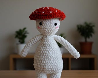 May include: A handmade crochet mushroom doll with a red cap dotted with white spots. The doll has a white body, arms, and legs, with black eyes and pink cheeks. The mushroom doll is standing on a wooden surface.
