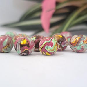 May include: A collection of round beads with a marbled effect. The beads are a mix of pink, green, yellow, and brown. The background is blurred, showing green leaves and a pink flower. The beads are likely for jewellery making.