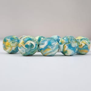 May include: A collection of marble-sized, spherical beads. Each bead features a swirling pattern of blue, yellow, and white, creating a marbled effect. The beads are arranged closely together, suggesting a craft or jewelry-making theme.