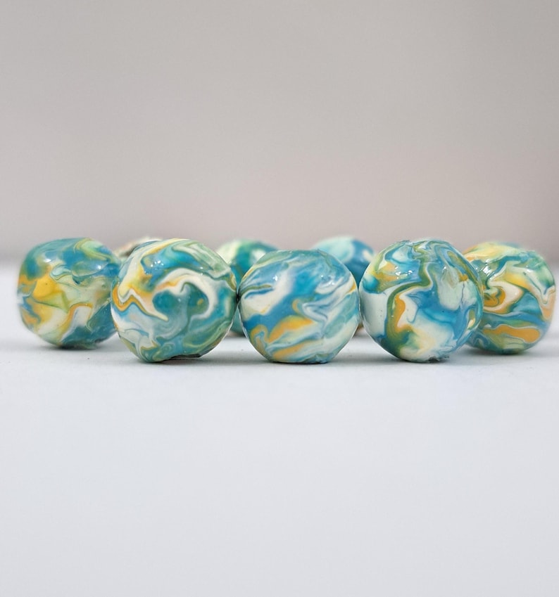 May include: A collection of marble-sized, spherical beads. Each bead features a swirling pattern of blue, yellow, and white, creating a marbled effect. The beads are arranged closely together, suggesting a craft or jewelry-making theme.