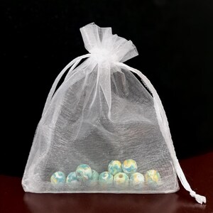 May include: A white, sheer organza drawstring bag filled with several round, light blue, yellow, and white beads. The bag is tied at the top with a matching white ribbon. The beads have a marbled appearance.