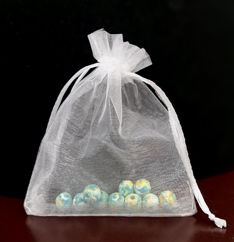 May include: A white, sheer organza drawstring bag filled with several round, light blue, yellow, and white beads. The bag is tied at the top with a matching white ribbon. The beads have a marbled appearance.