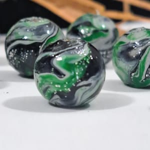 May include: Close-up of several glass marbles with a swirling design. The marbles feature a mix of black, green, and silver colors. The marbles are round and have a glossy finish. The marbles are on a white surface.