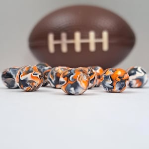 May include: A collection of round beads with a marbled design in orange, black, and white. A brown football is in the background. The beads are arranged in a group, with a focus on their unique patterns and colors.