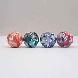 Puede incluir: Cuatro cuentas esféricas con patrones arremolinados. Las cuentas muestran una variedad de colores, incluyendo rojo, blanco y amarillo; verde, negro y blanco; azul y blanco; y rojo, gris y blanco. Las cuentas están dispuestas en una fila sobre un fondo blanco.