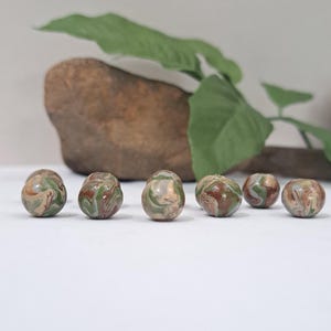 May include: Six small, rounded, decorative stones with a marbled appearance. The stones feature swirls of green, brown, and cream colors. A large rock and green leaf are in the background.
