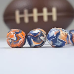 May include: Close-up of several small, round beads with a marbled design in shades of blue, orange, and white. A blurred brown American football with white stitching is in the background. The beads are the focal point.