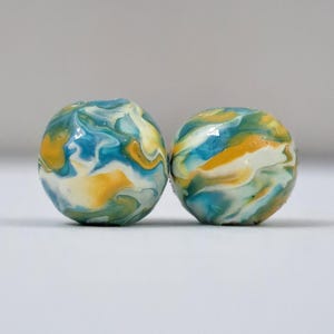 May include: Two round, marbled beads. The beads feature a swirl of colors, including blue, yellow, green, and white. The beads are smooth and glossy, with a unique pattern. The background is a plain, light gray.