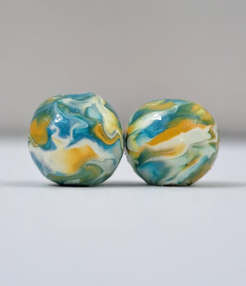 May include: Two round, marbled beads. The beads feature a swirl of colors, including blue, yellow, green, and white. The beads are smooth and glossy, with a unique pattern. The background is a plain, light gray.