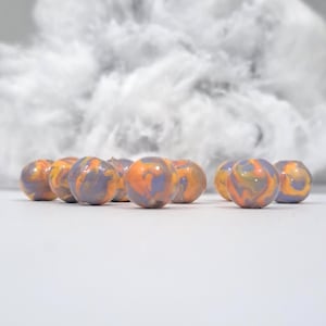Puede incluir: Un conjunto de esferas con aspecto de mármol, con un diseño de remolino en naranja, morado y amarillo. Se colocan frente a un fondo de material blanco y esponjoso, creando un contraste de texturas y colores.