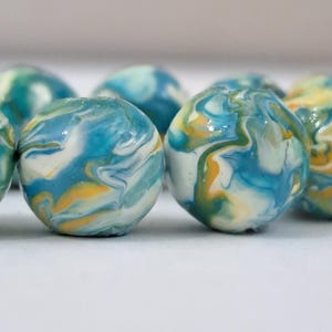 May include: Close-up of several round beads with a marbled effect. The beads feature swirls of blue, white, and yellow, creating a unique, artistic appearance. The beads are arranged on a white surface.