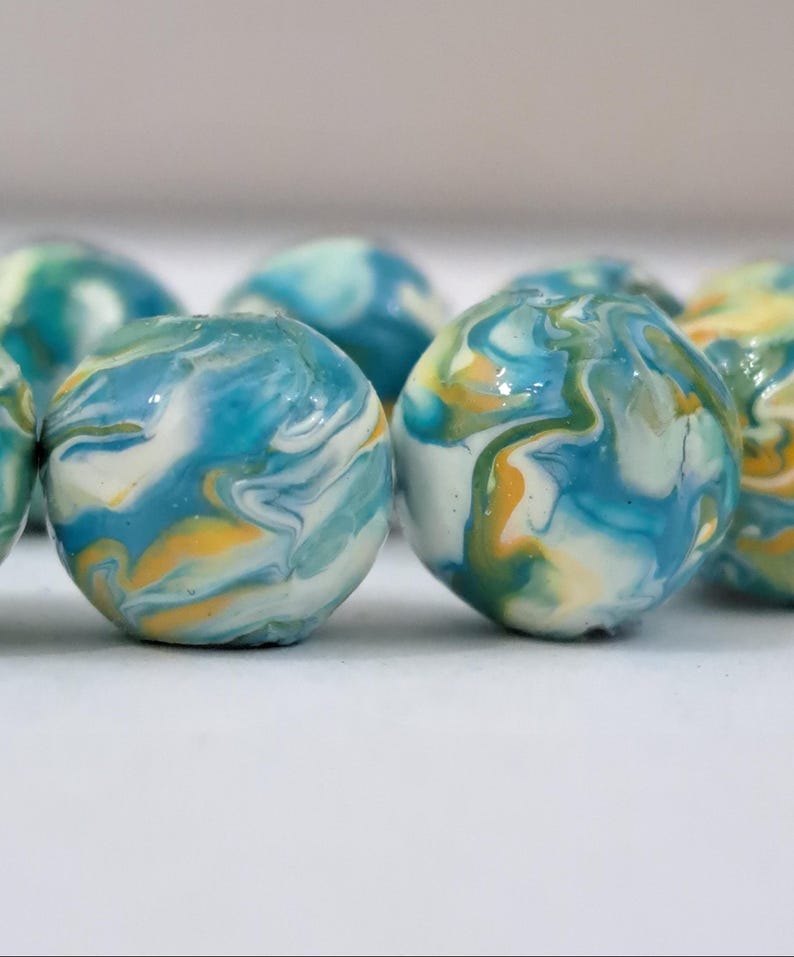 May include: Close-up of several round beads with a marbled effect. The beads feature swirls of blue, white, and yellow, creating a unique, artistic appearance. The beads are arranged on a white surface.