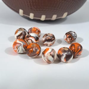 May include: A collection of marbled beads in shades of orange, brown, white, and black. The beads are spherical and have a swirled pattern. A brown football is in the background.