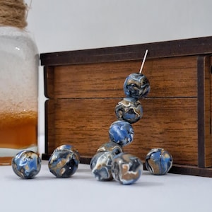 May include: A collection of blue, gold, and black marbled beads. Some beads are strung on a white pin, while others are scattered on a white surface. A wooden box and a glass bottle are in the background.