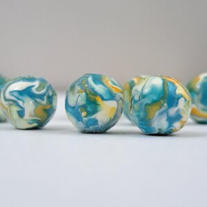 May include: Several small, round beads with a marbled appearance. The beads feature swirls of blue, white, and yellow colors. The beads are arranged on a white surface, with a soft focus background.