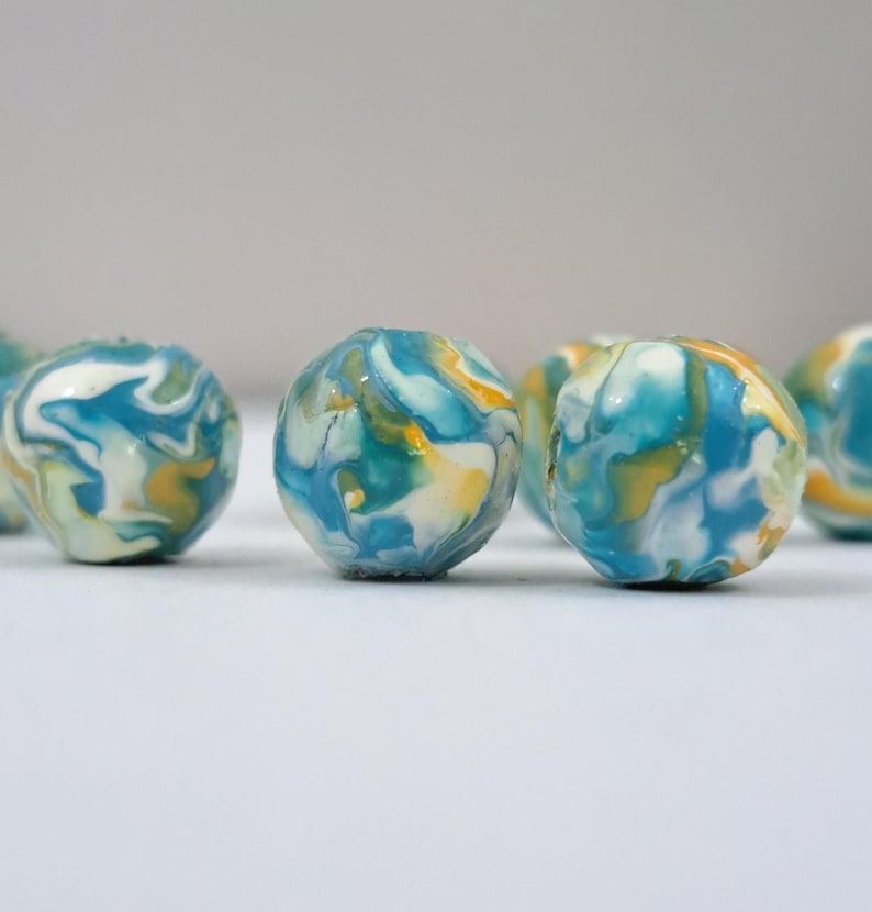 May include: Several small, round beads with a marbled appearance. The beads feature swirls of blue, white, and yellow colors. The beads are arranged on a white surface, with a soft focus background.