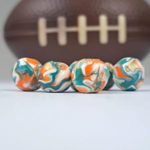 May include: Six colorful, swirled beads in shades of orange, teal, and white are in the foreground. A brown football with white stripes is blurred in the background. The beads have a marbled appearance.