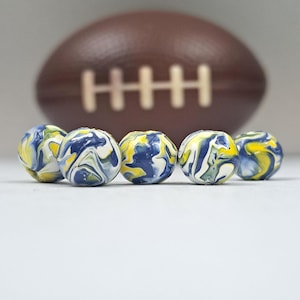 Könnte beinhalten: Fünf kleine, runde Murmeln mit einem Wirbelmuster aus Blau, Gelb und Weiß im Vordergrund. Ein brauner Football mit weißen Nähten ist im Hintergrund unscharf. Die Murmeln sind wahrscheinlich für ein Spiel oder ein Bastelprojekt.