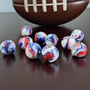 May include: A collection of small, round beads with a marbled design in red, white, and blue. A brown football is in the background. The beads are scattered on a dark surface.