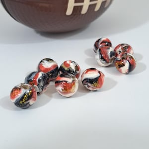 Op de afbeelding: Een verzameling decoratieve kralen met een gemarmerd ontwerp in rood, zwart, wit en goud. De kralen zijn rond en hebben een glanzende afwerking. Op de achtergrond bevindt zich een bruin object in de vorm van een American football.
