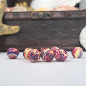May include: A collection of colorful, marbled beads in shades of red, purple, and yellow. The beads are spherical and appear to be made of a smooth material. A wooden chest and seashells are in the background.