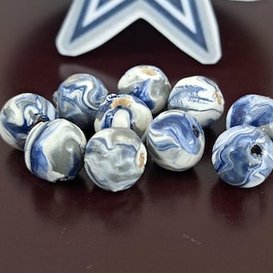 May include: Ten spherical beads with a marbled pattern in blue, grey, and white. The beads have a glossy finish and are arranged on a dark brown surface. A star-shaped decoration is visible in the background.