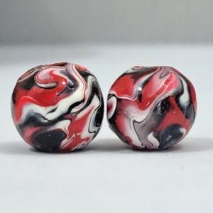 Puede incluir: Dos cuentas esféricas con un patrón jaspeado en colores rojo, negro, blanco y gris. La superficie de cada cuenta es lisa y brillante, mostrando un diseño arremolinado y multicolor que mezcla los distintos tonos en un movimiento fluido.